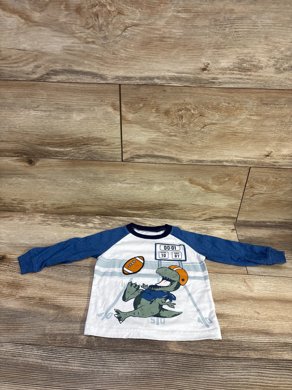 Front view of Carter’s football shirt in white with navy sleeves, featuring an alligator football graphic on a wood floor