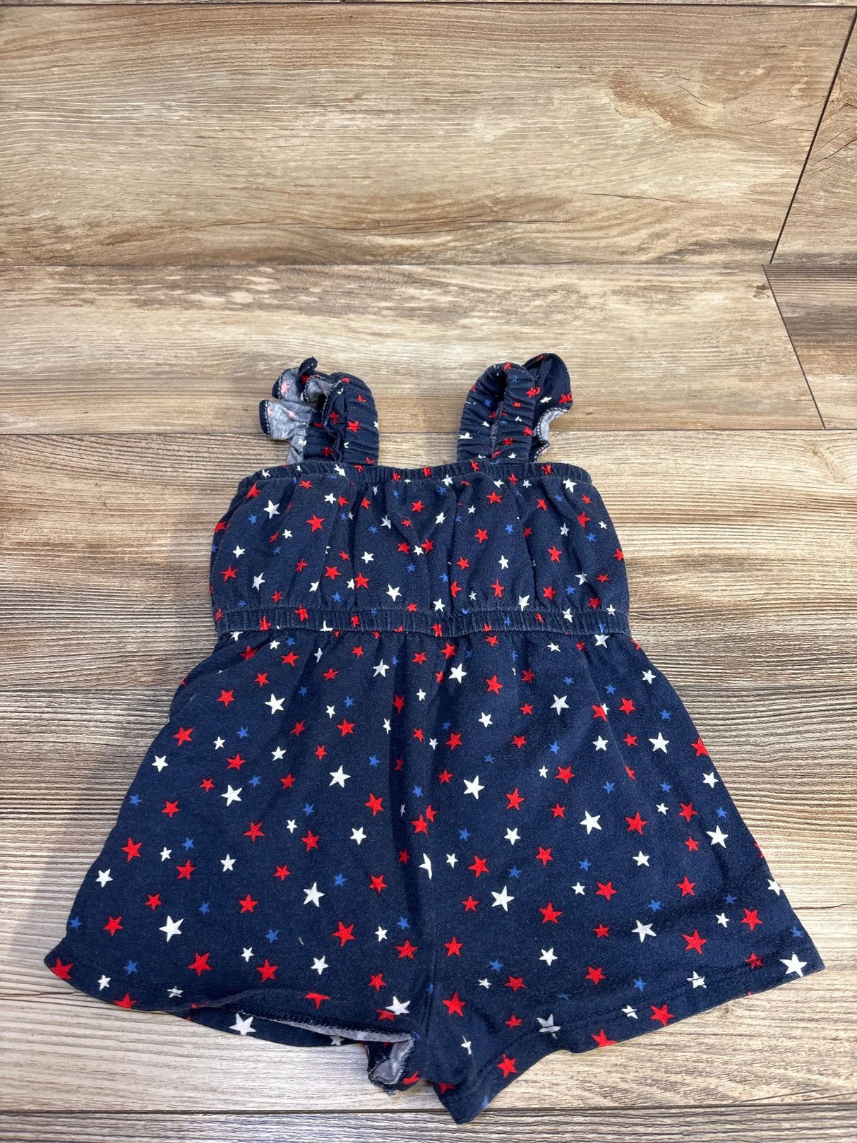 Flat-lay front of navy toddler sleeveless romper (size 12-18 months) showcasing red, white, and blue star print, ruffle shoulder straps, and elasticized bodice on a light wood floor
