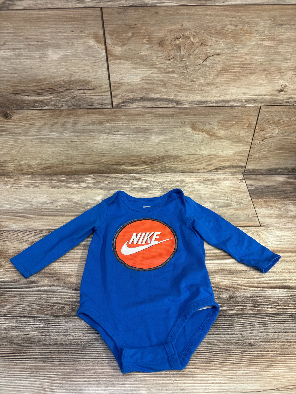 Top view of a blue Nike long sleeve bodysuit in size 9m showcasing its orange circular logo on a wood-look floor