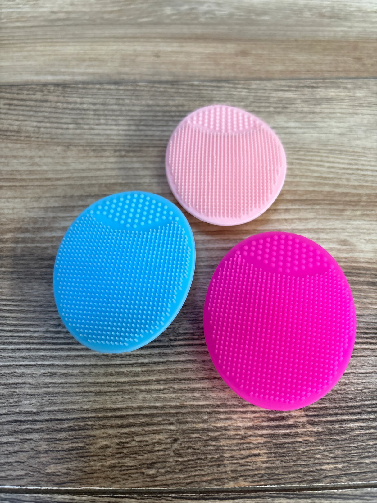Three colorful silicone face scrubbers in blue, pink, and magenta on wood surface