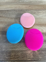 Three colorful silicone face scrubbers in blue, pink, and magenta on wood surface