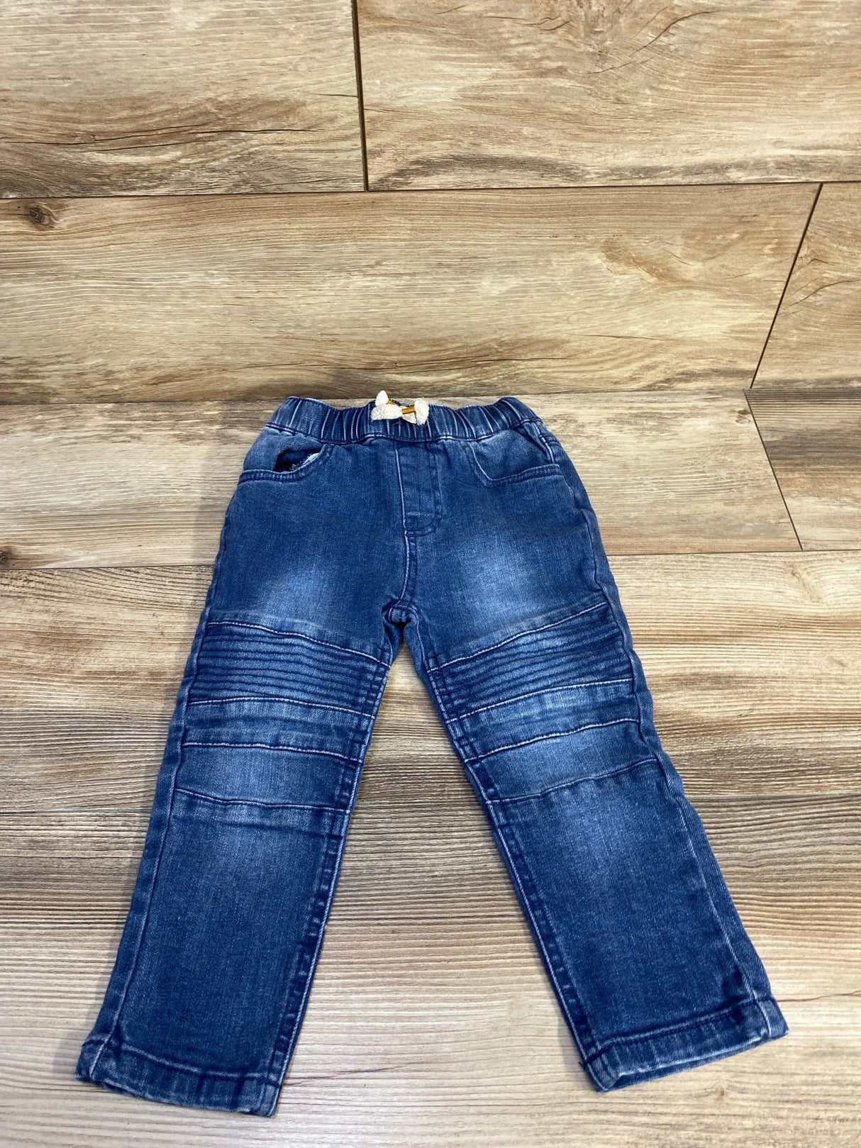 Flat lay of Art Class blue toddler drawstring jeans showcasing elastic drawstring waistband, stitched knee ribbing and faded wash on wood floor