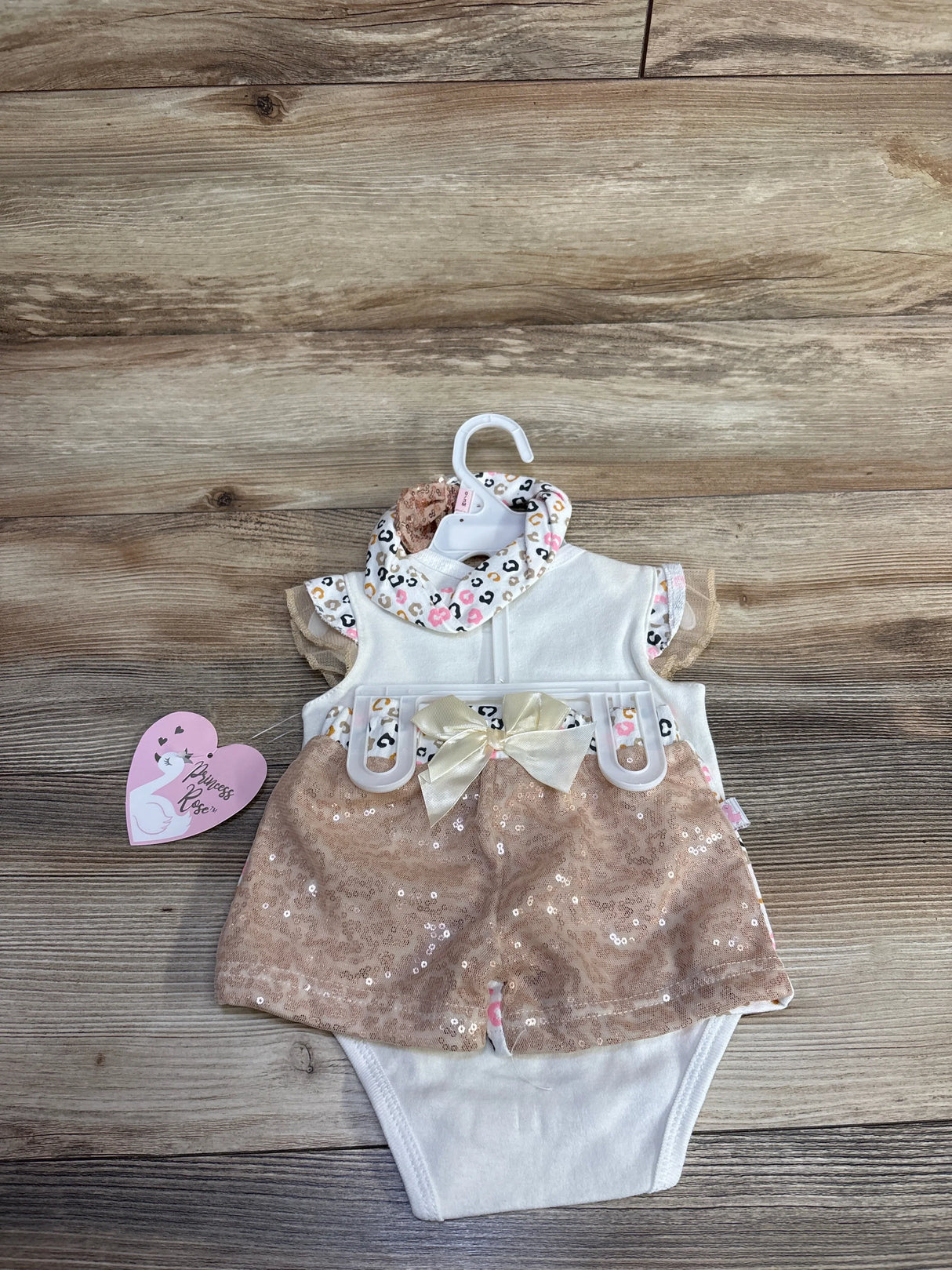 Top view of white baby bodysuit with rose pink sequin shorts, satin bow and leopard print collar, matching headband on wood