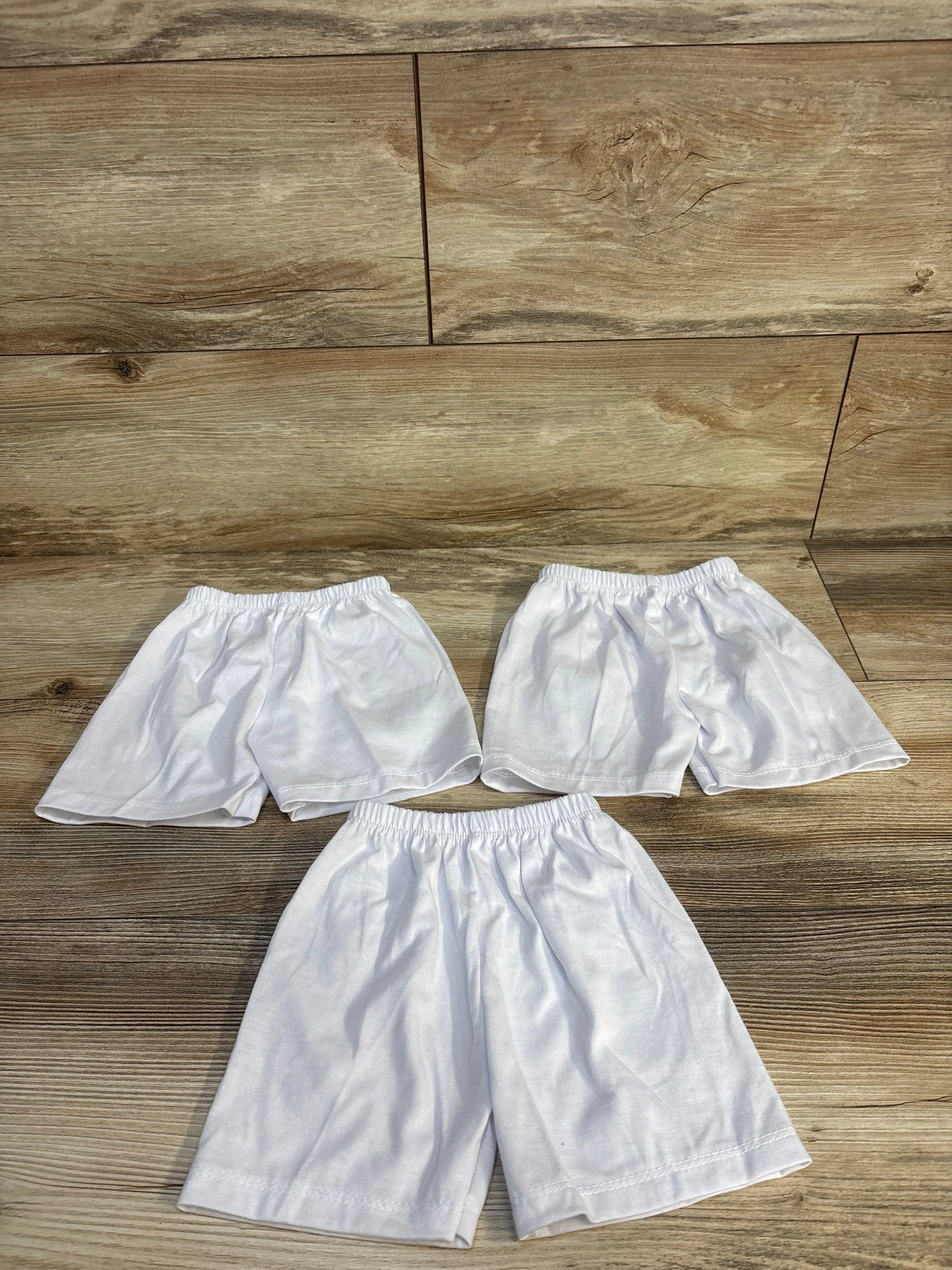 Three pairs of white kids' shorts with elastic waistbands on wood floor, ideal for baby and toddler clothing resale