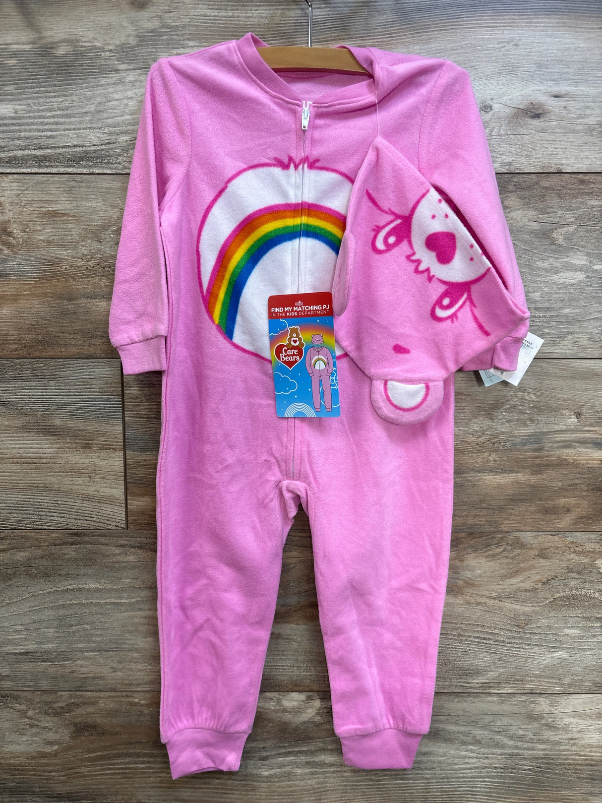 Front view of pink Care Bears zip sleeper with rainbow belly graphic and matching hat showing bear face on wood background