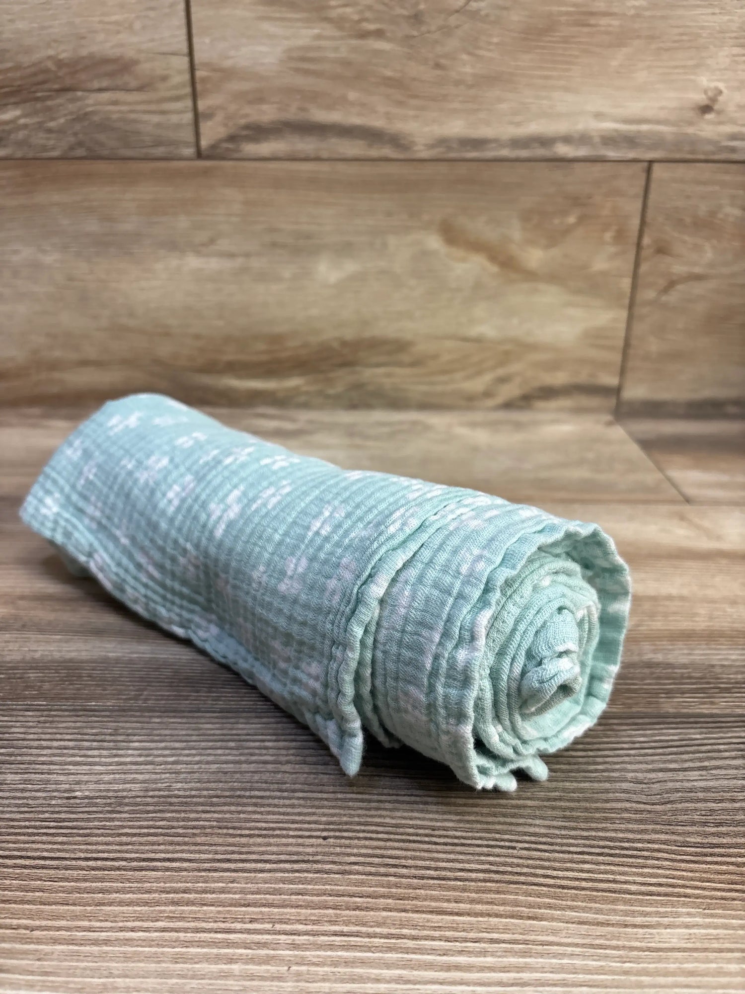 Angled view of an Aden and Anais blue muslin swaddle blanket, 44 by 44 inches, rolled on a wood floor