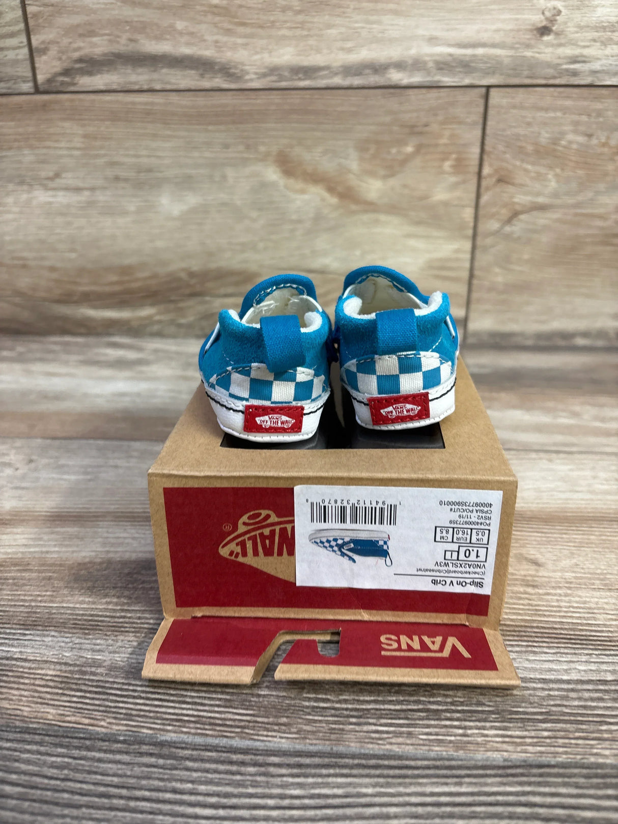 Rear view of baby Vans Slip-On crib shoes in blue checkered canvas showing red 'Off The Wall' heel tags on a Vans box on wood floor