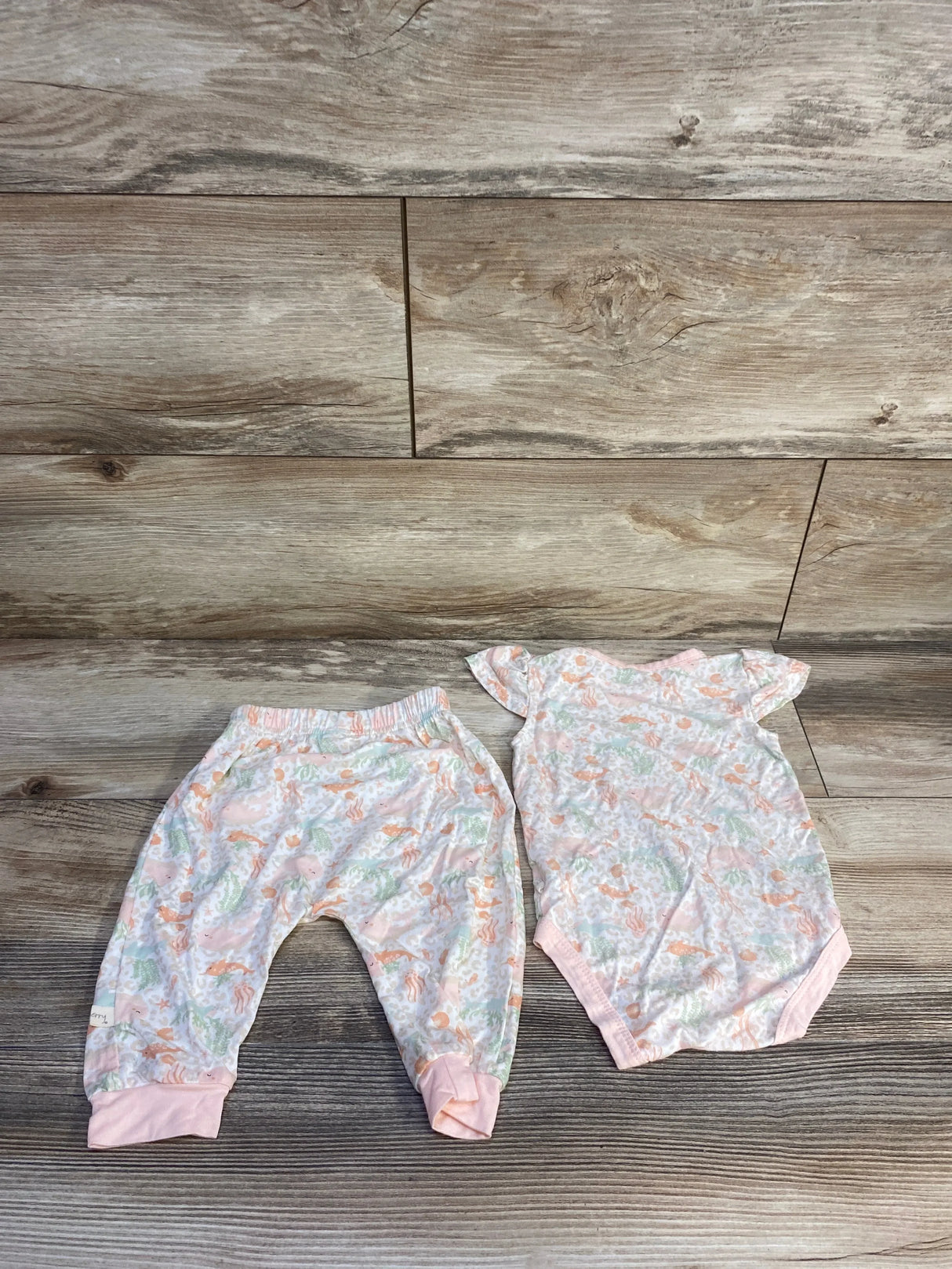 Flat lay of Milkberry two-piece sea animal bodysuit and pants with pastel print, pink trim and elastic waist on wood background