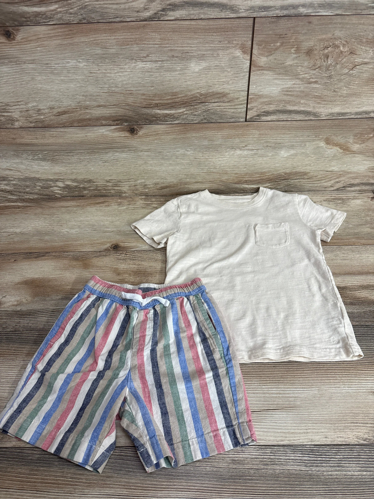 Flat lay of boys' cream slub pocket tee and linen-cotton multicolor striped shorts showing front view on wood floor