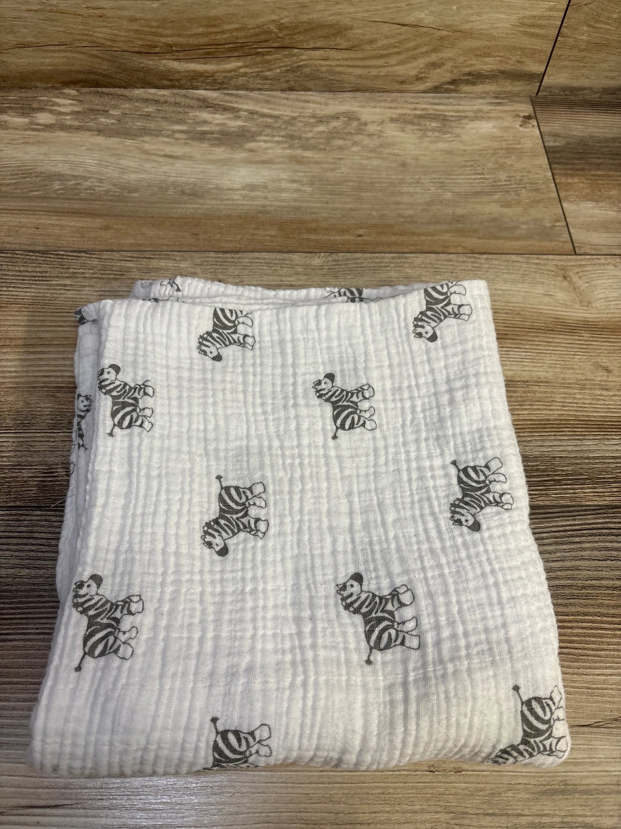 Top view of Amazing Baby muslin swaddle blanket in white with gray zebra print, laid flat on a wood floor