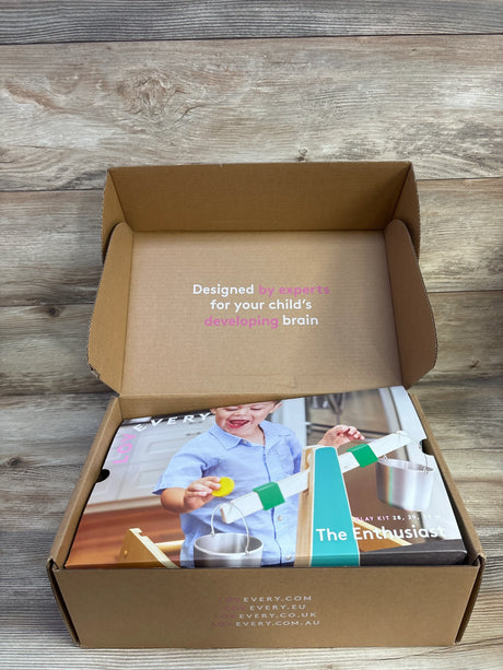 Open box with Lovevery Enthusiast Play Kit showing lid text 'Designed by experts for your child's developing brain' on wood