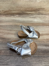 Metallic silver toddler sandals with bow detail and cushioned soles on wood background