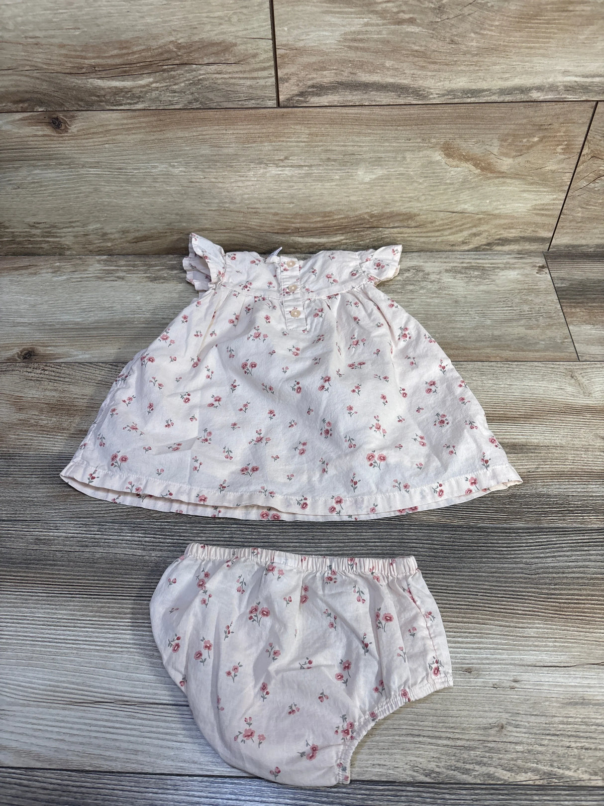 Flat lay of pink floral baby dress and matching bloomers showcasing ruffle cap sleeves, front buttons, and elastic waist on wood background