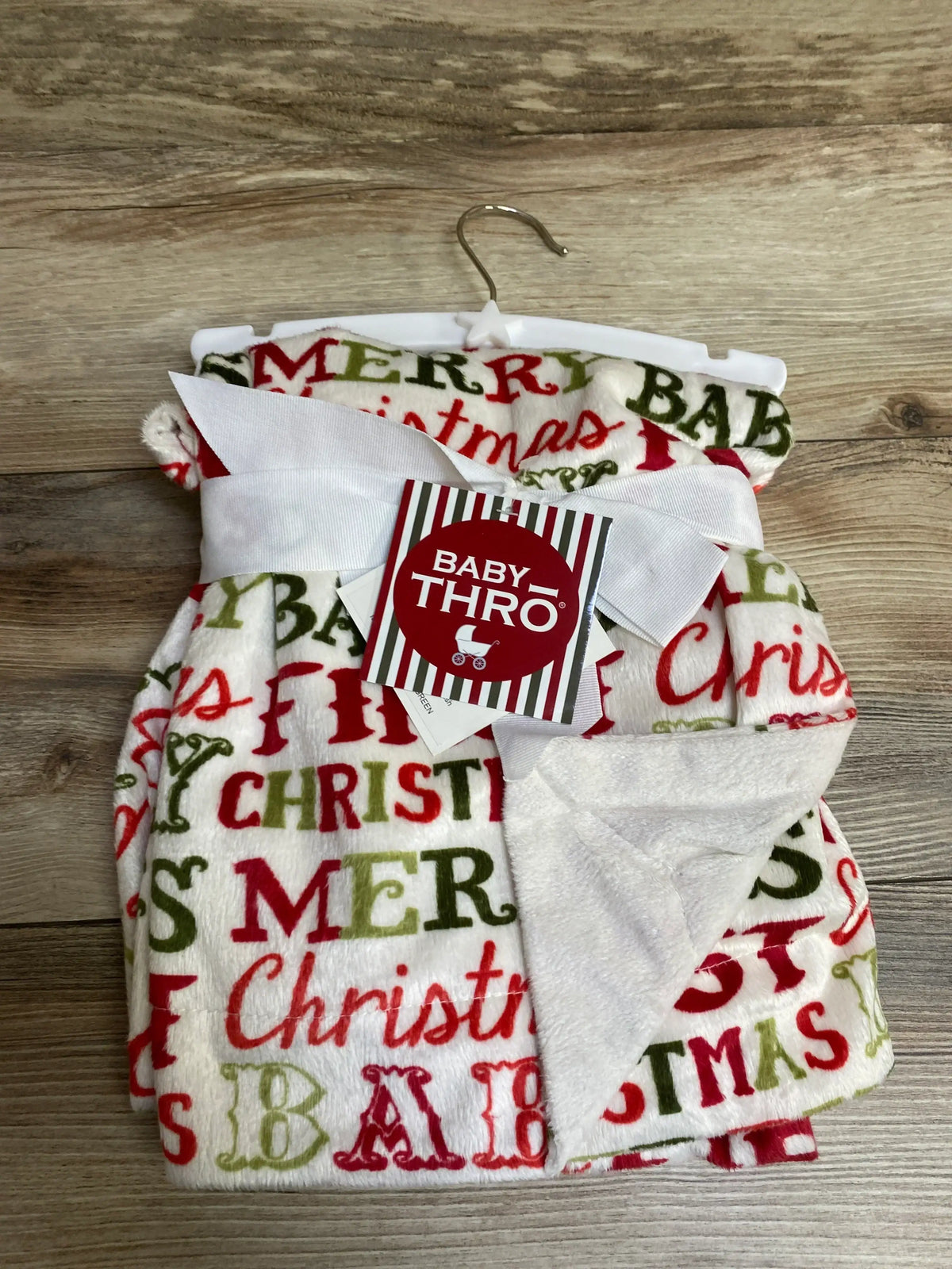 Baby Thro Merry Christmas holiday blanket with red and green text, soft fleece, white ribbon, on hanger