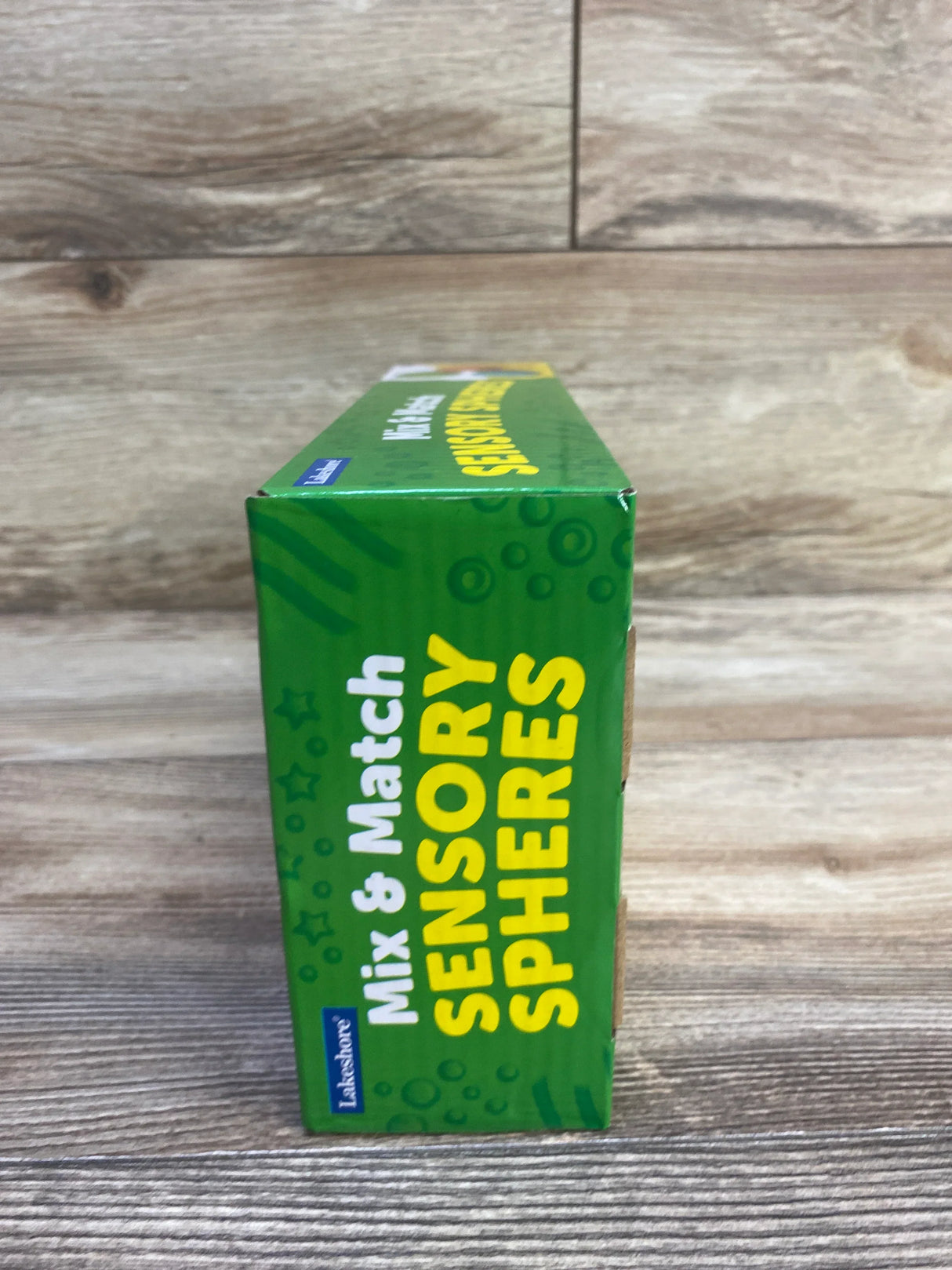 Side view of Lakeshore Mix & Match Sensory Spheres box showing green packaging with large yellow 'Sensory Spheres' text on wood background