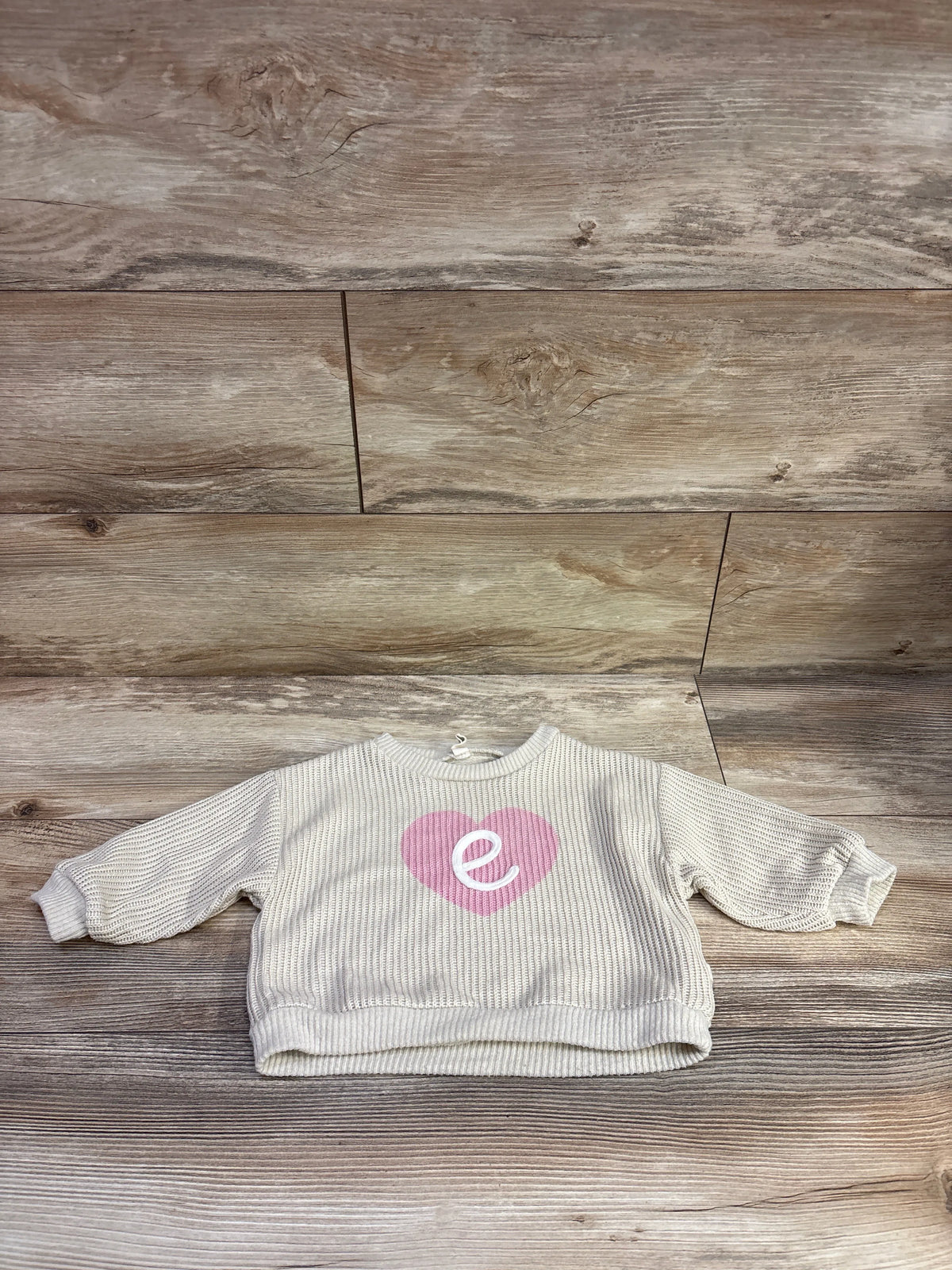 Front view of beige knit initial E sweater showcasing ribbed texture and pink heart detail on a wood background