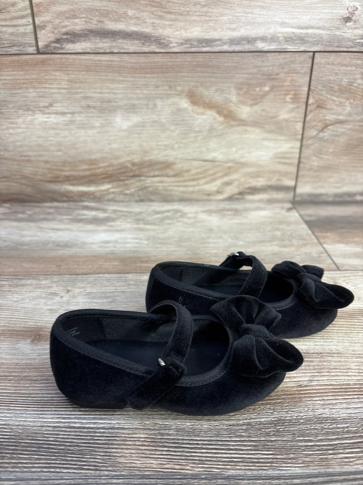 Angled view of Baby Gap black velvet Mary Jane flats showcasing front bow and adjustable strap on wood floor background