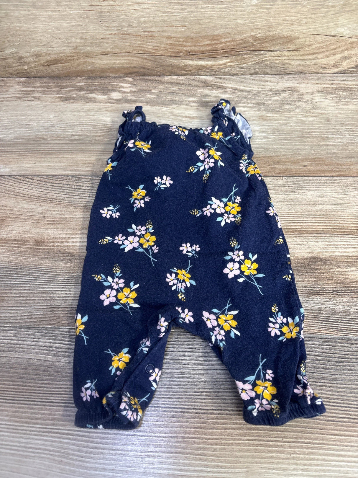 Flat lay of navy floral newborn sleeveless jumpsuit with ruffle straps, snap leg closures and elastic cuffs on wood background