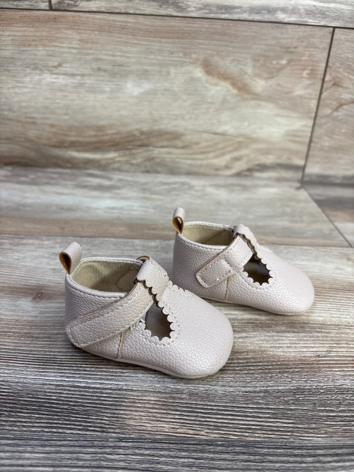 Three-quarter view of beige soft-sole baby shoes with scalloped T-strap and pebbled leather on light wood background