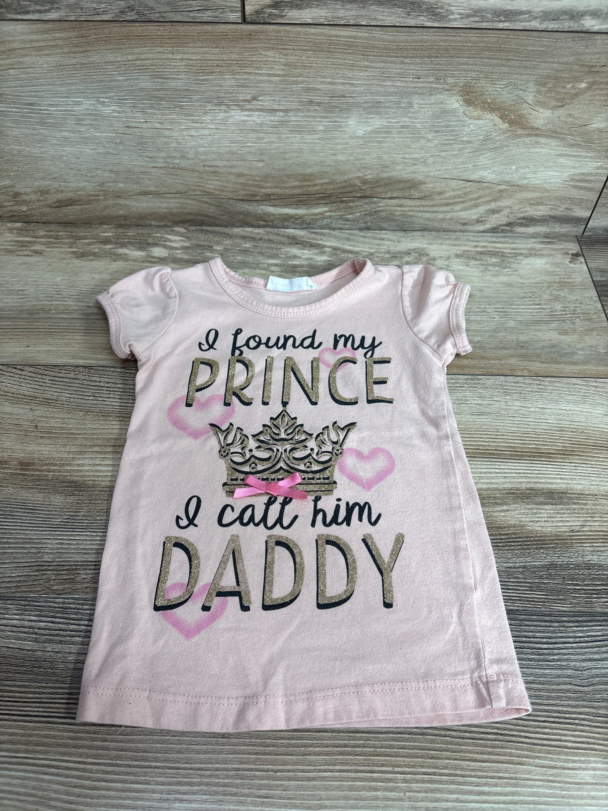 Flat-lay of pink girls' 3T short-sleeve t-shirt showcasing glitter text I found my prince I call him Daddy with crown and hearts on wood background