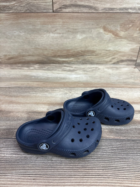 Angled view of navy Crocs Classic Clogs showcasing ventilation holes and heel straps on a wood-grain background