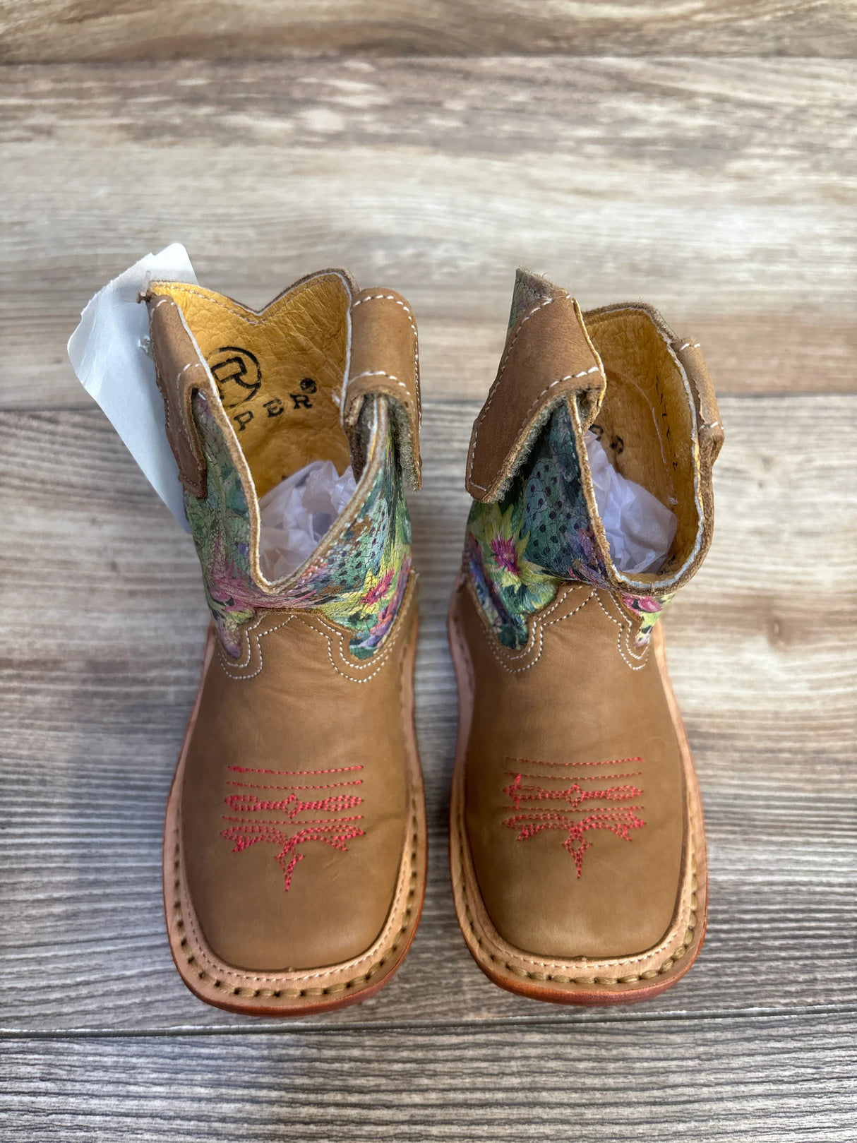 Kids brown leather cowboy boots with floral embroidery and pink stitching, on wood floor