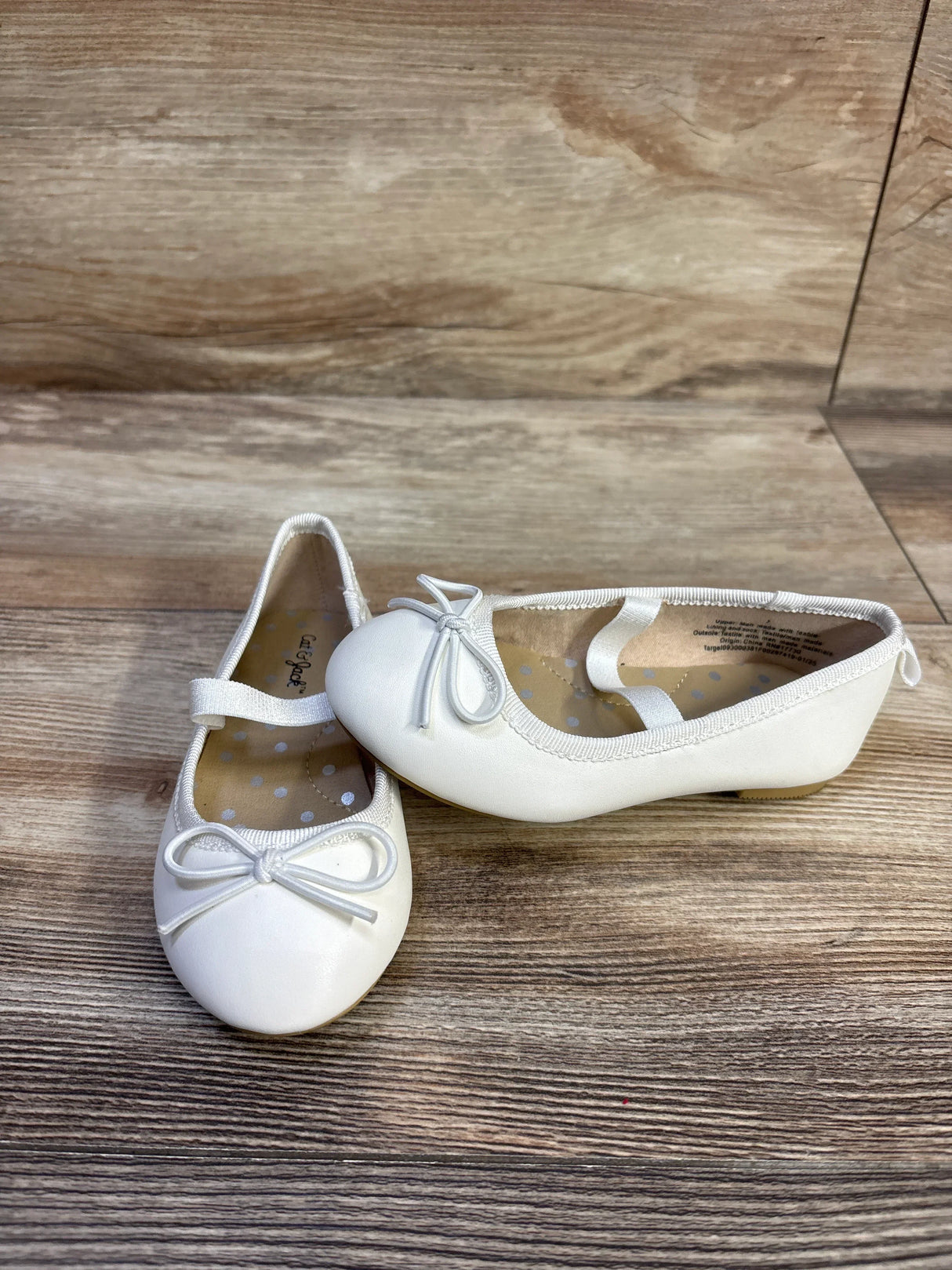 Angled front view of Cat & Jack Nora white slip-on ballet flats showing bow, elastic strap and polka-dot insole on wood floor