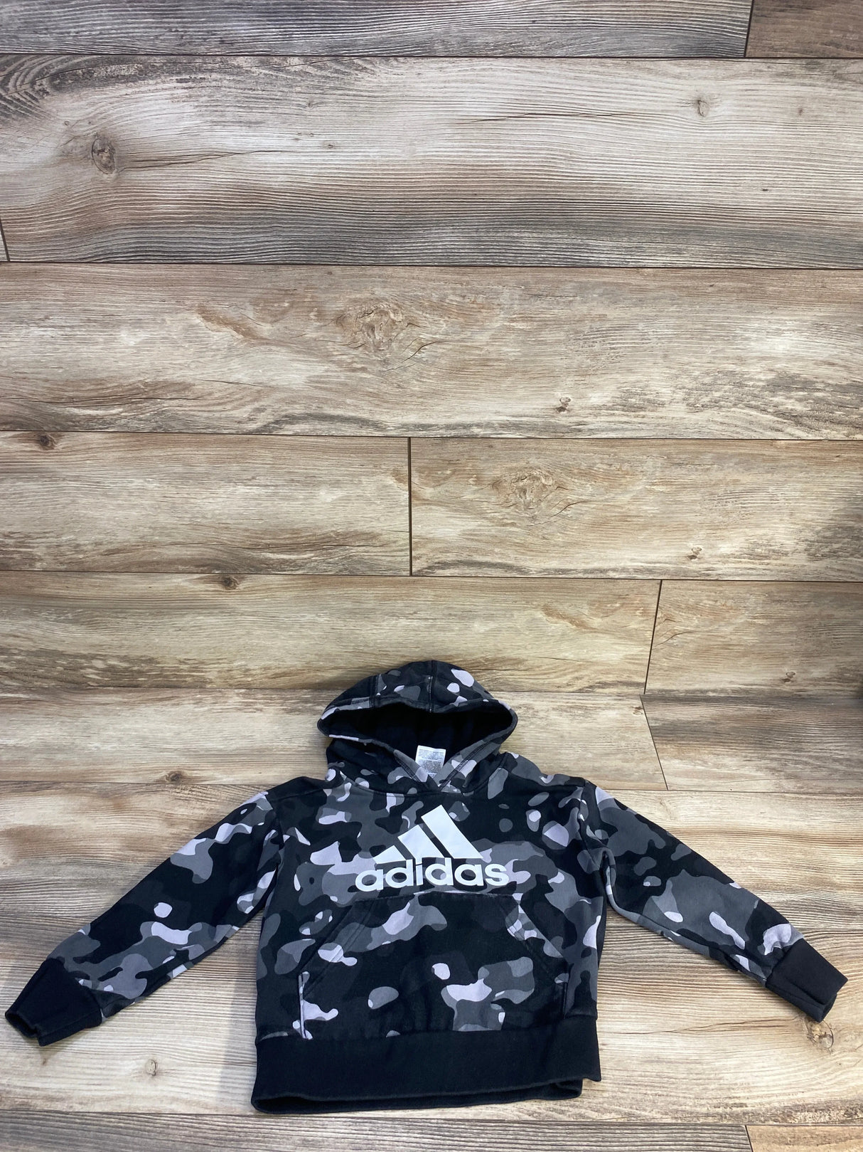 Front view of toddler Adidas pullover sweatshirt in grey camo showcasing white adidas logo, hood, and kangaroo pocket on wood floor background