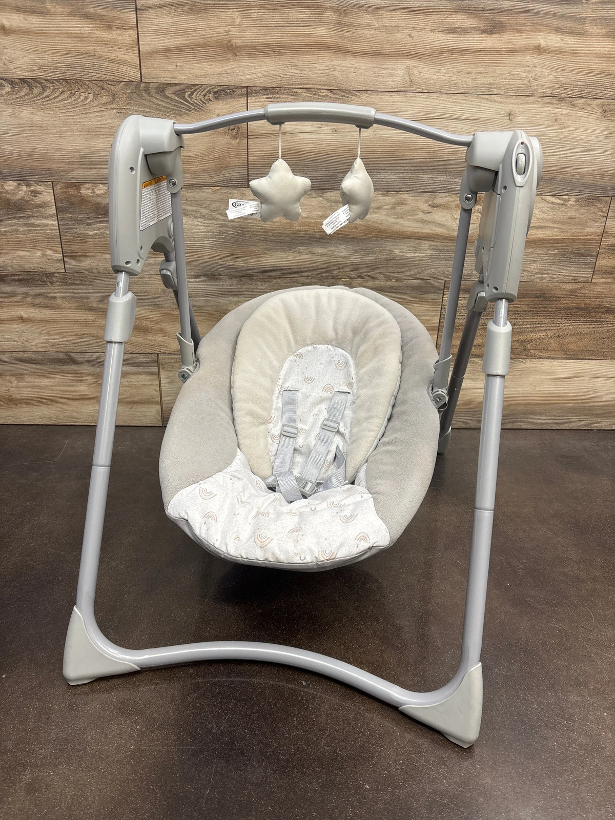 Front view of Graco Slim Spaces Compact Baby Swing in Reign with padded seat, safety harness, and hanging plush toys