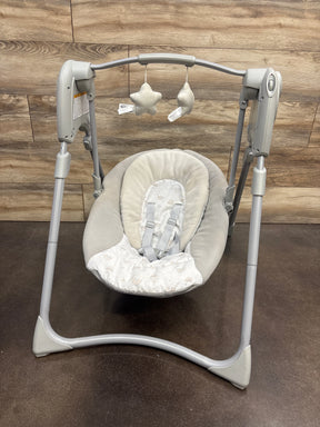 Front view of Graco Slim Spaces Compact Baby Swing in Reign with padded seat, safety harness, and hanging plush toys