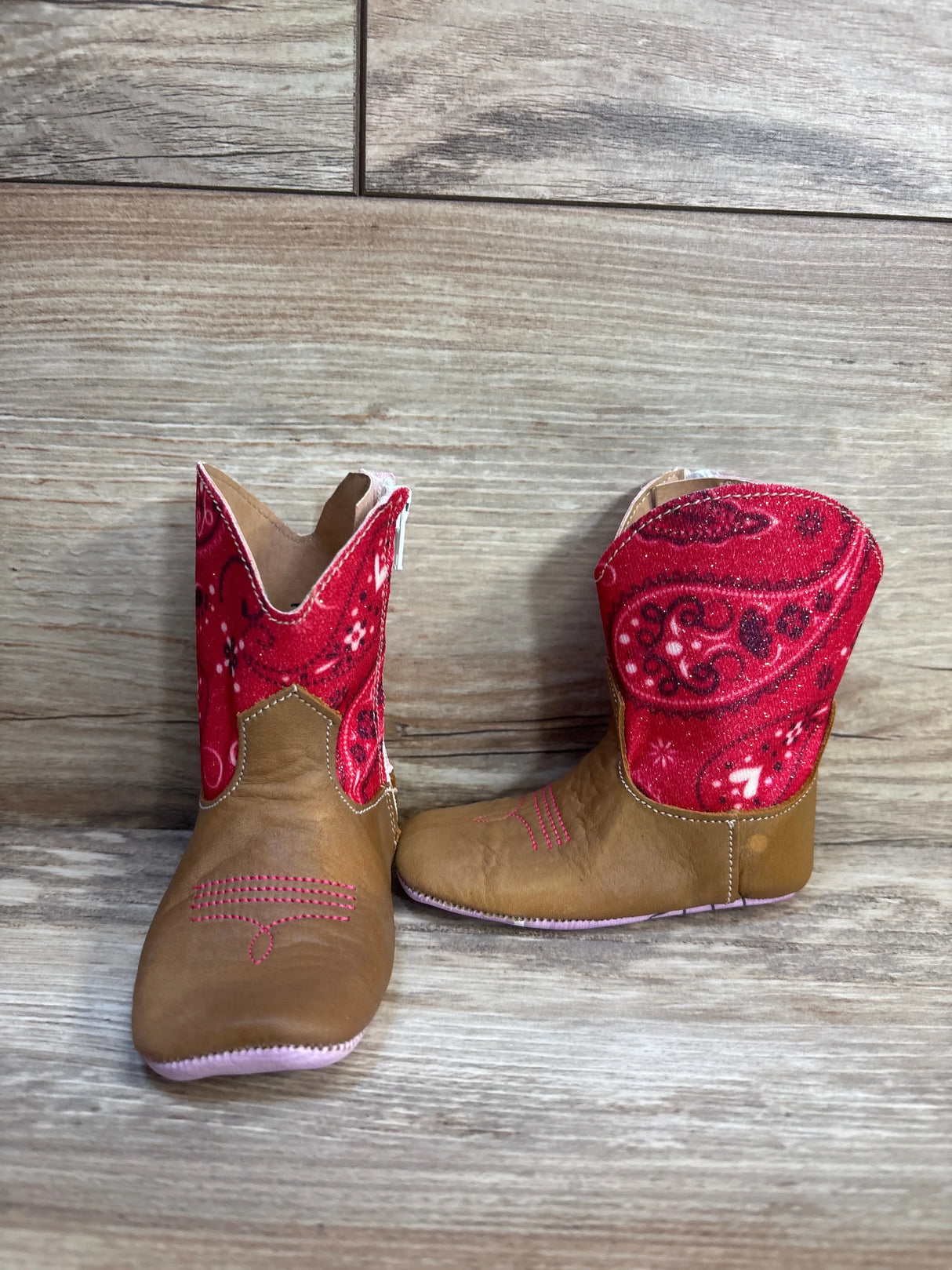 Front and side view of Tin Haul mini square-toe toddler boots showing brown faux-leather foot, red sparkly paisley shaft and soft pink sole on wood background