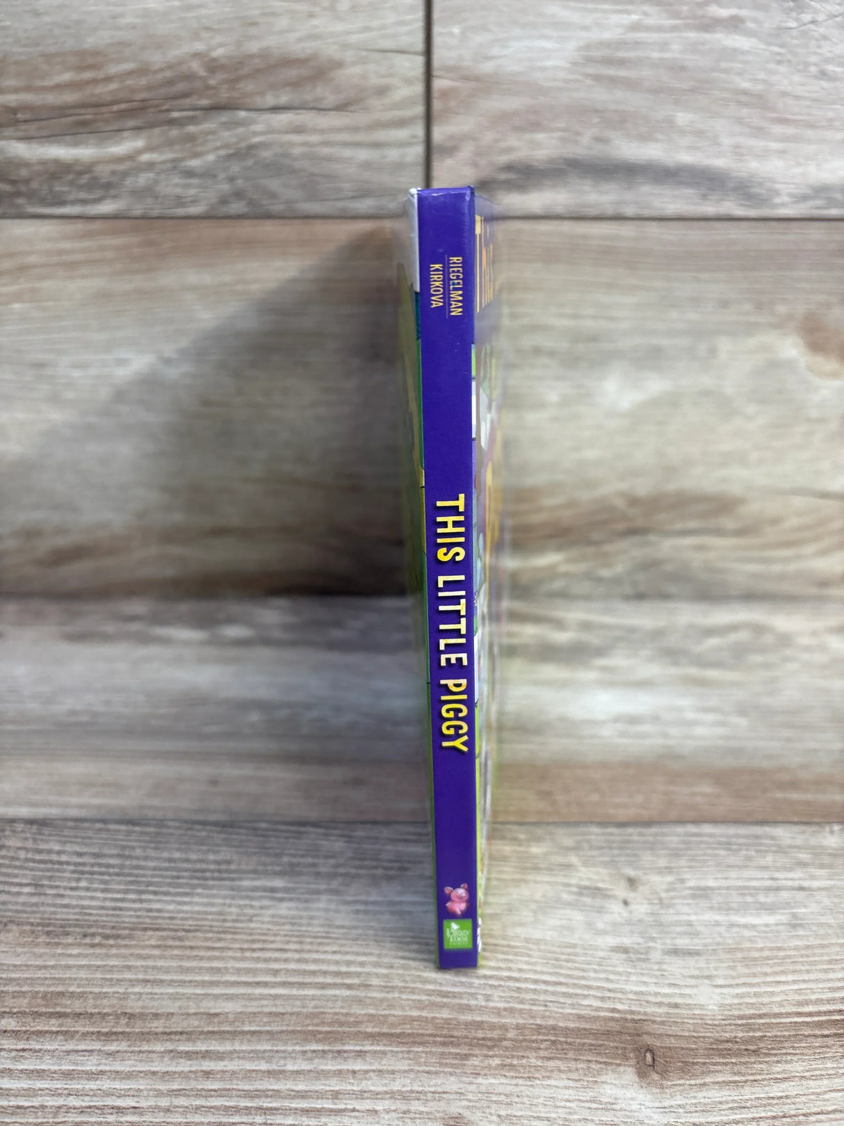 Spine view of This Little Piggy board book showing purple spine with vertical title and small pig icon on wood background