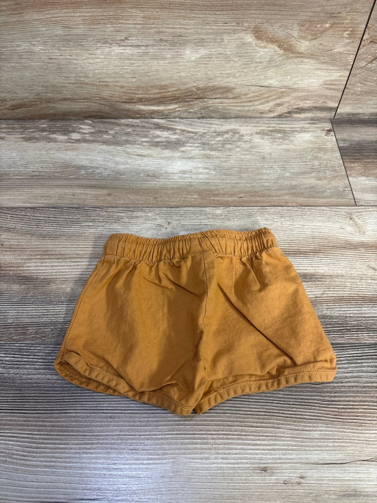 Flat lay of Little Co. organic baby shorts in caramel showing elastic waistband and rolled hem on wood background, size 12 months