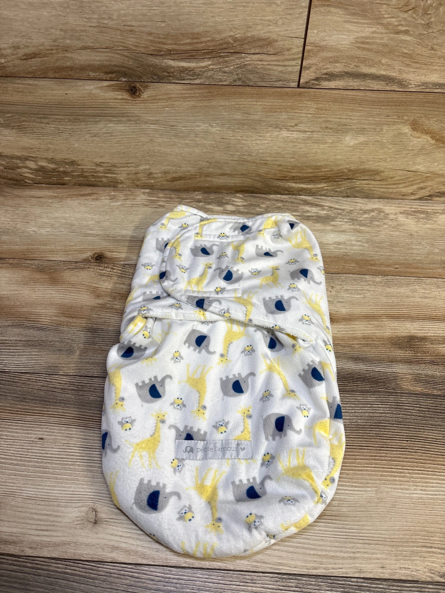 Top view of Petite L’amour plush swaddle wrap showcasing white plush fabric with giraffe, elephant, and bee print on a wood background