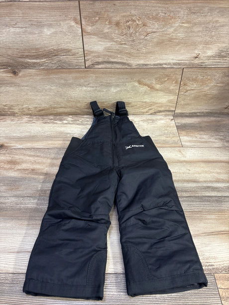 Front view of black Arctix insulated snow bib overalls showcasing adjustable shoulder straps and knee panels on a wood floor