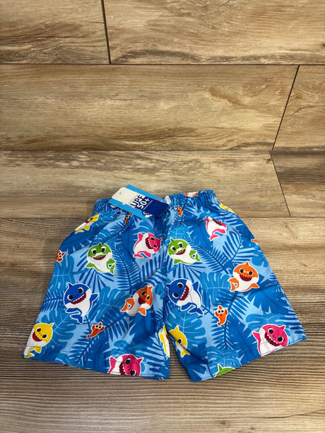 Top-down view of blue toddler swim trunks with elastic waistband and Baby Shark characters over tropical leaf print, tag showing UPF 50 plus on wood floor