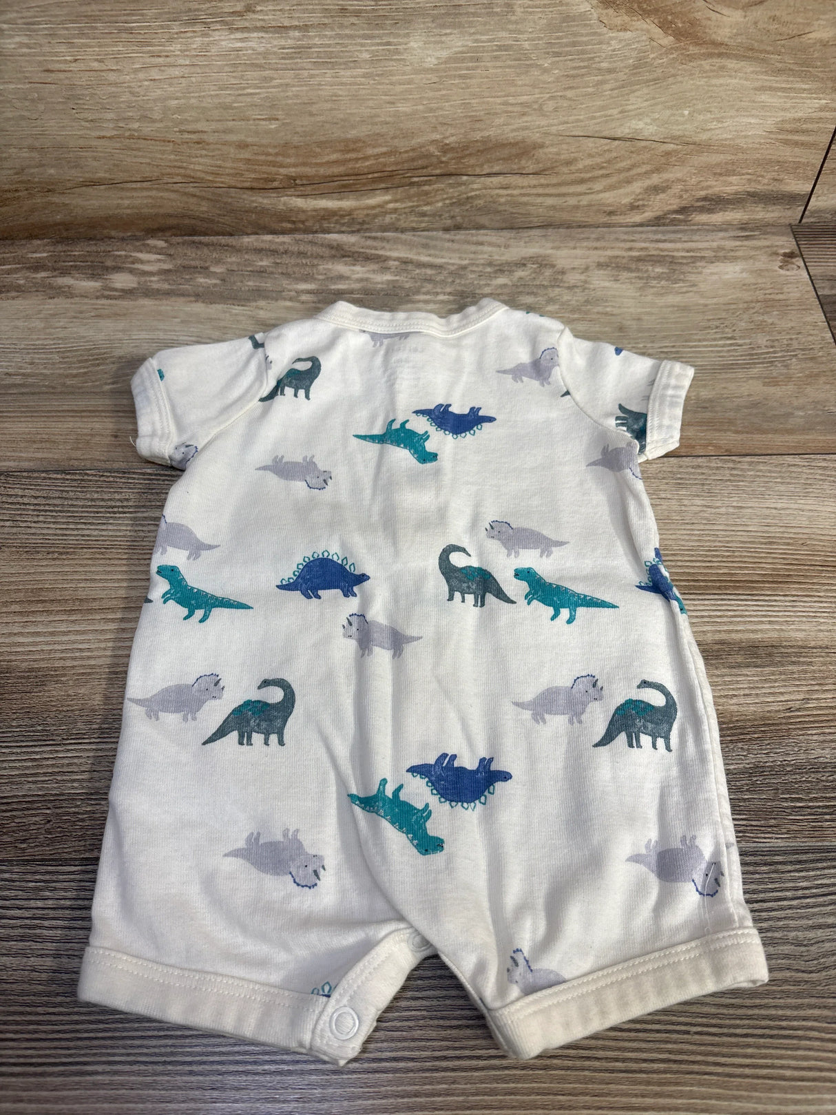 Flat lay of ivory button-front shortie romper size 3M showing blue and teal dinosaur print on wood background