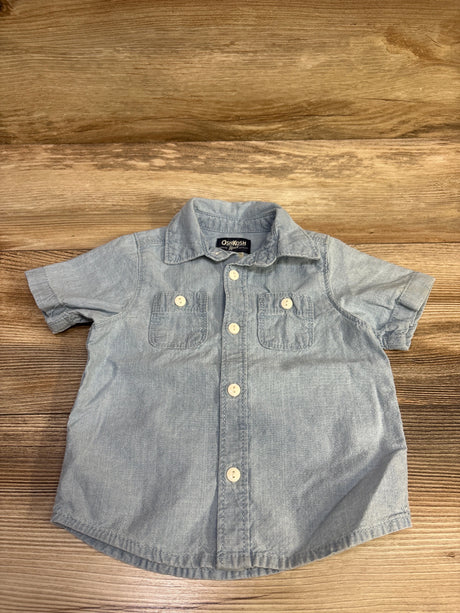 Front view of OshKosh blue chambray button-up shirt with short sleeves, collar, buttons, and two chest pockets on wood grain