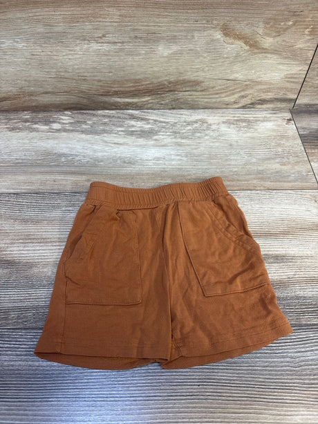 Top-down view of caramel brown toddler shorts showing elastic waistband and two front patch pockets on wood-look background