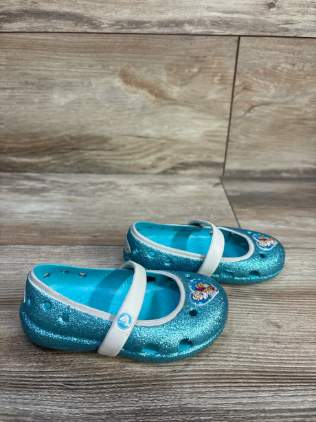 Angled front view of children's glitter blue Crocs Mary Jane flats showing white strap and Frozen character charm on wood floor