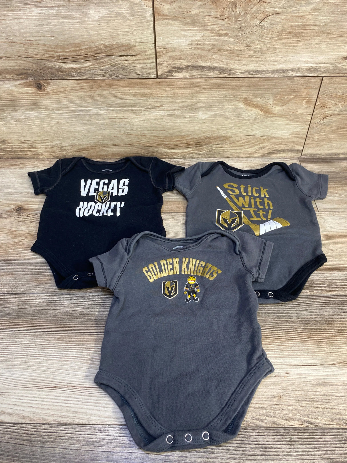 Flat lay of three baby bodysuits showing Vegas Golden Knights logos and prints on grey and black fabric on wood background