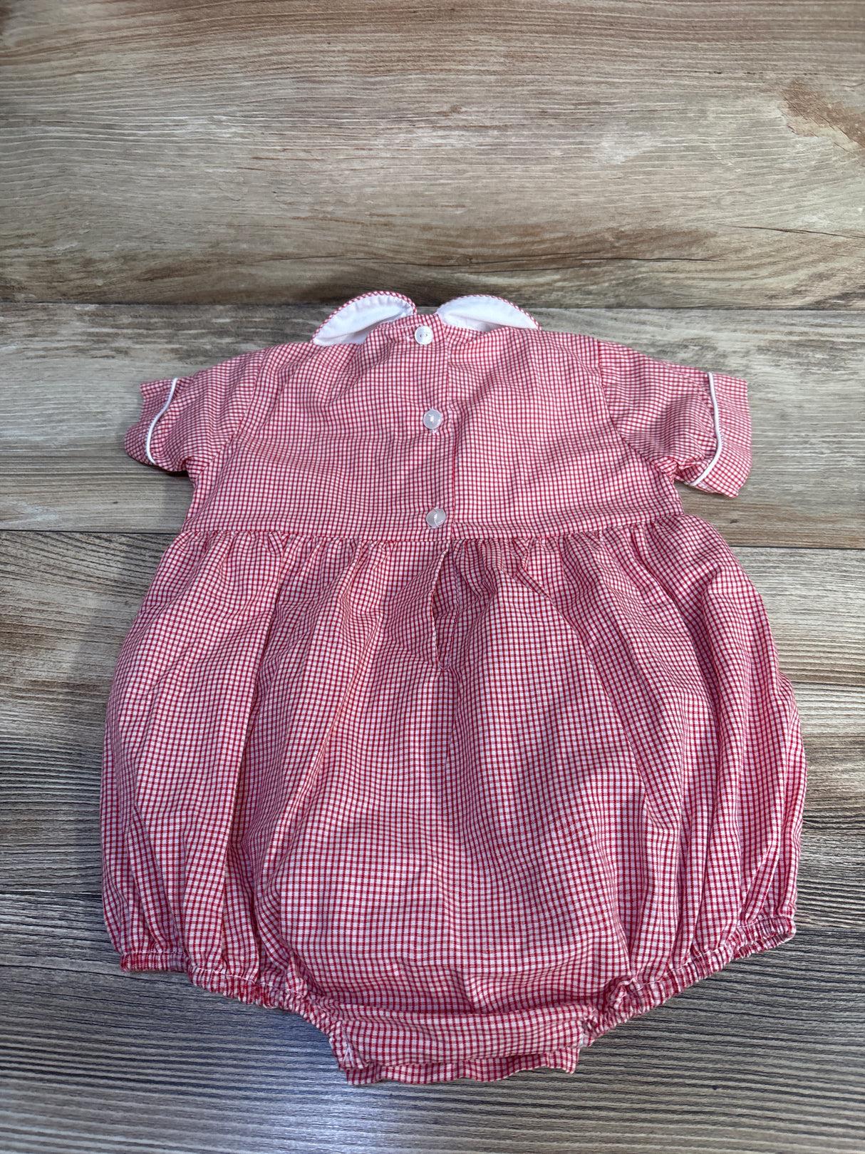 Top-down view of red gingham baby bubble romper with white scalloped Peter Pan collar, back buttons, and elastic leg openings on wood background