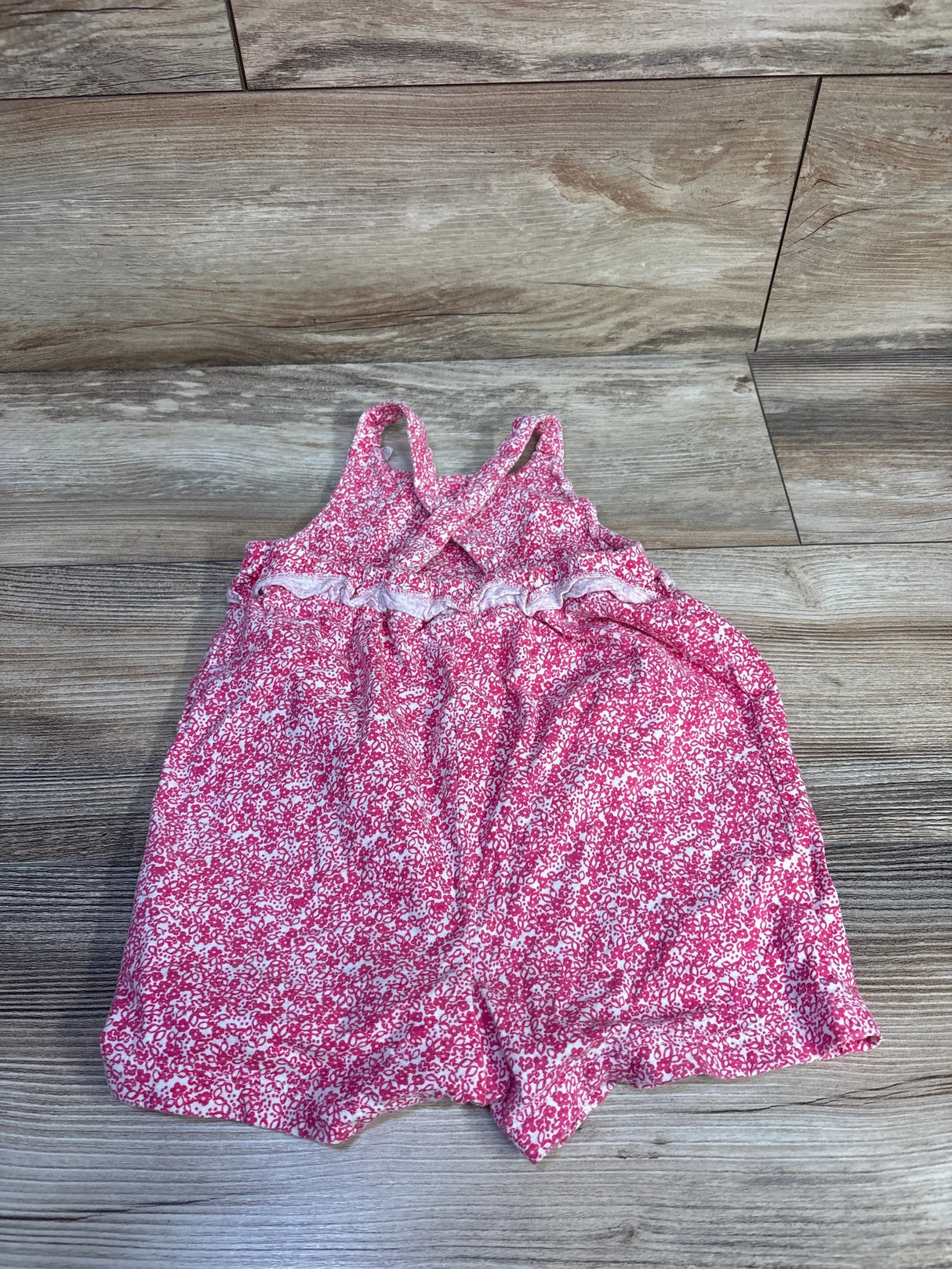 Flat lay of pink floral tank romper showing crossed back straps and ruffle waist on light wood floor