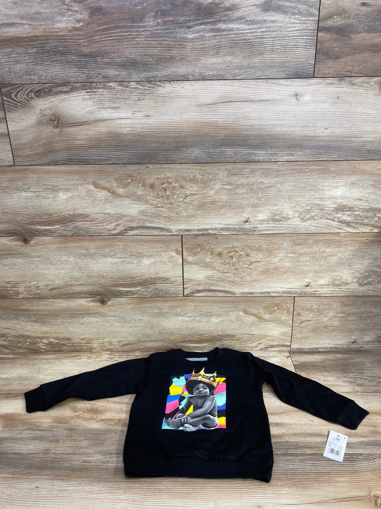Black kids crewneck sweatshirt with colorful graphic of a baby wearing a gold crown, displayed on wood floor