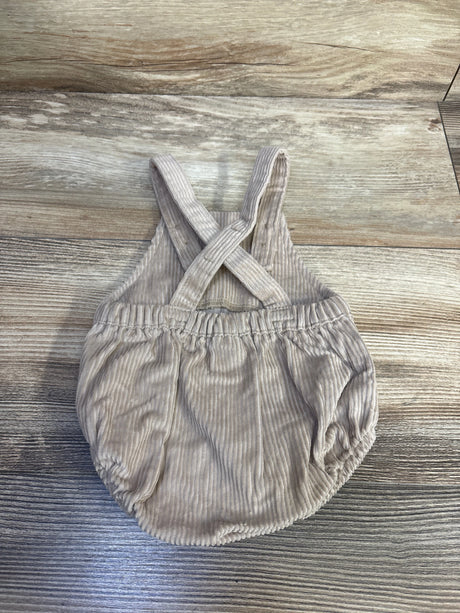 Top-down view of beige newborn corduroy shortalls showcasing crossed back straps and elasticized waist and leg openings on a wood background