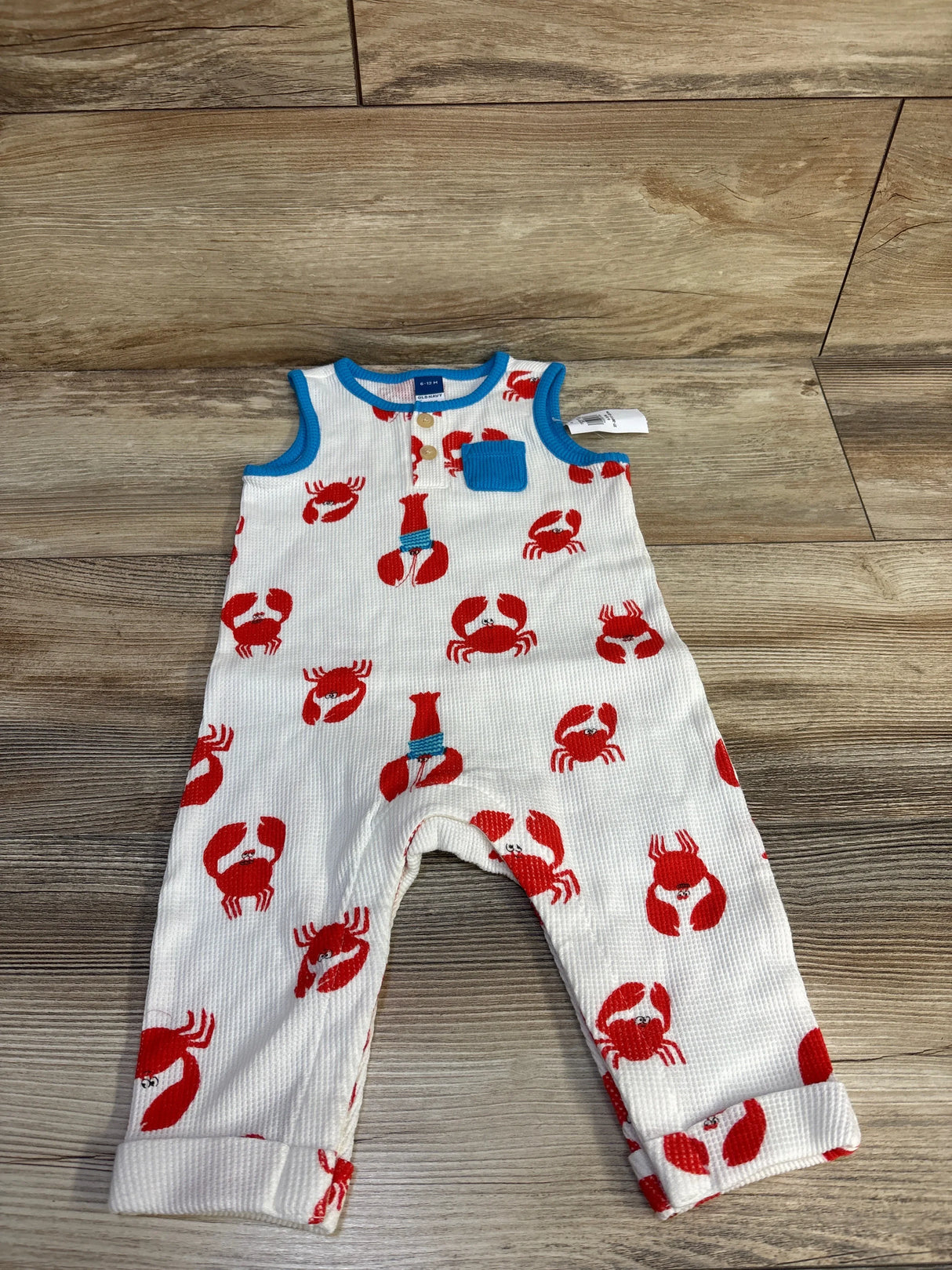 Top-down view of baby sleeveless waffle-knit Henley jumpsuit showcasing red crab print, blue trim and chest pocket on wood background