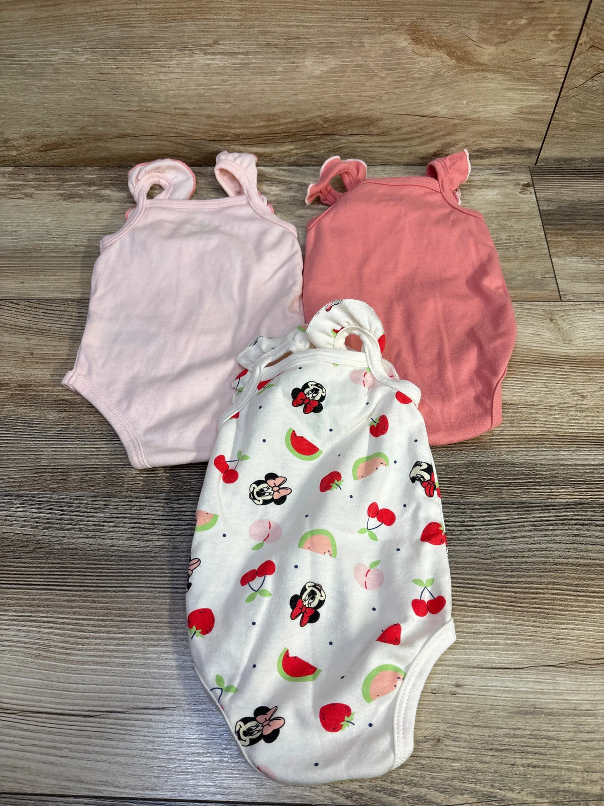 Flat lay of three sleeveless Disney Minnie Mouse baby bodysuits: one white with Minnie and fruit print and two solid pink on wood background