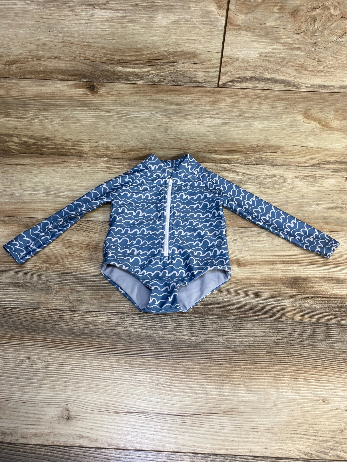 Blue baby rash guard with white wave print, long sleeves, front zipper, laid on wood floor