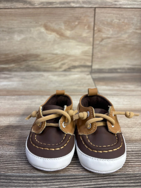 Front view of Sperry boat crib shoes in brown and tan showing white toe caps, contrast stitching and knotted laces on a wood grain surface