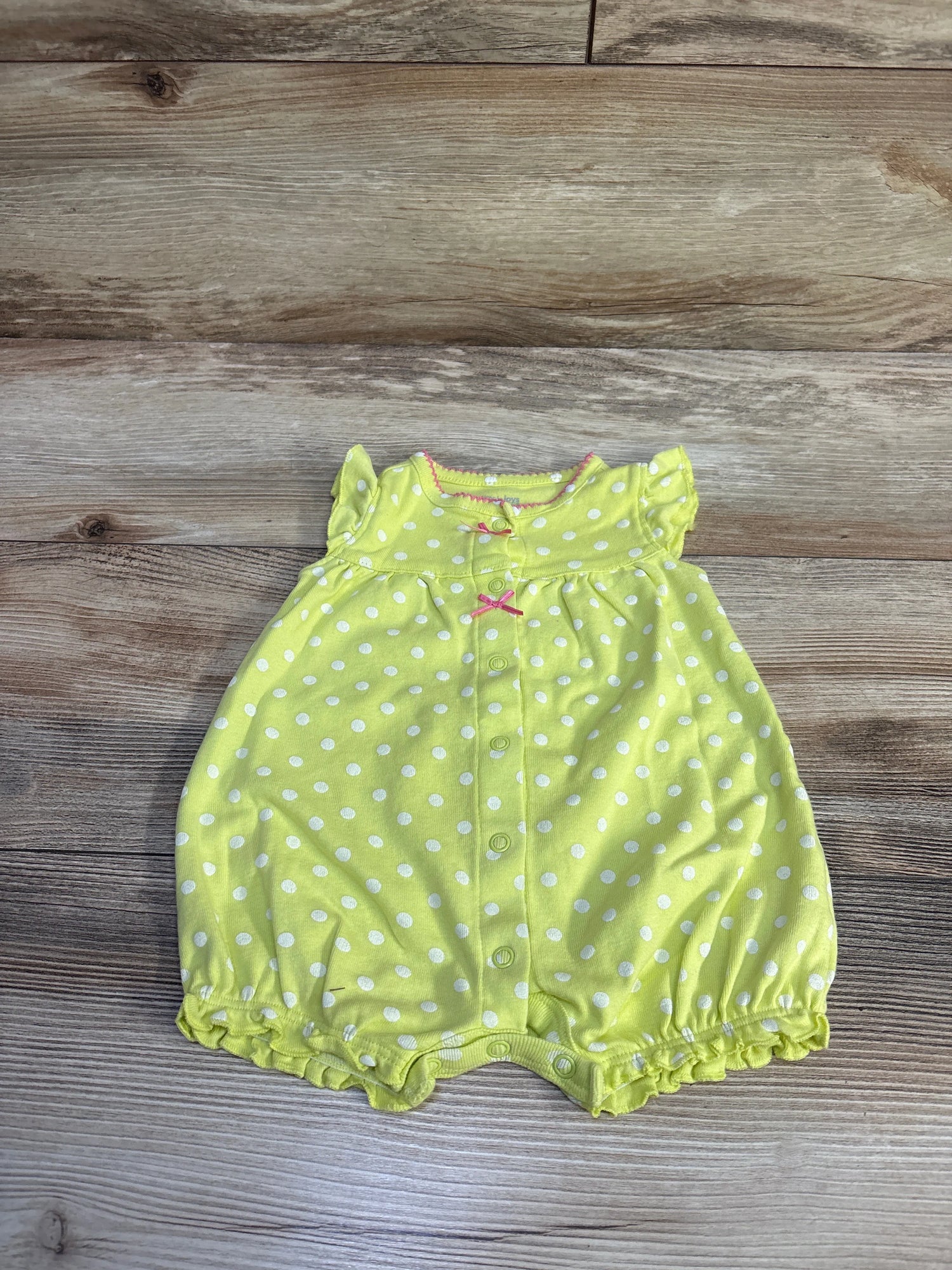 Flat lay of lime polka dot baby shortie romper with front snap buttons, pink bow accents and ruffled sleeves on wood background
